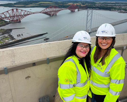 Amy and Lyndsay from CHAS in white hard hats and hi-vis jackets at the top of the Queensferry Crossing with the Forth Road and Rail bridges in the background