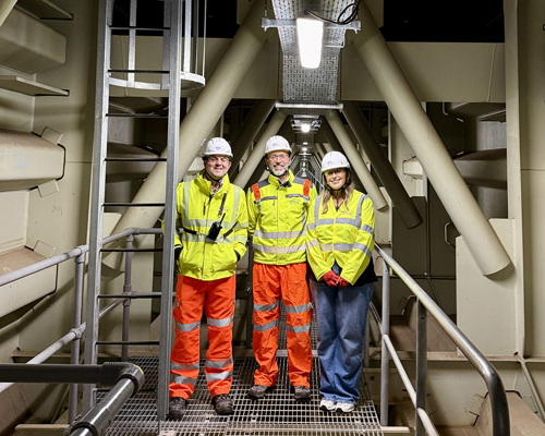 Three people smiling at the camara in hard hats and hi-vis gear staning on an enclosed walkway in the Queensferry Crossing